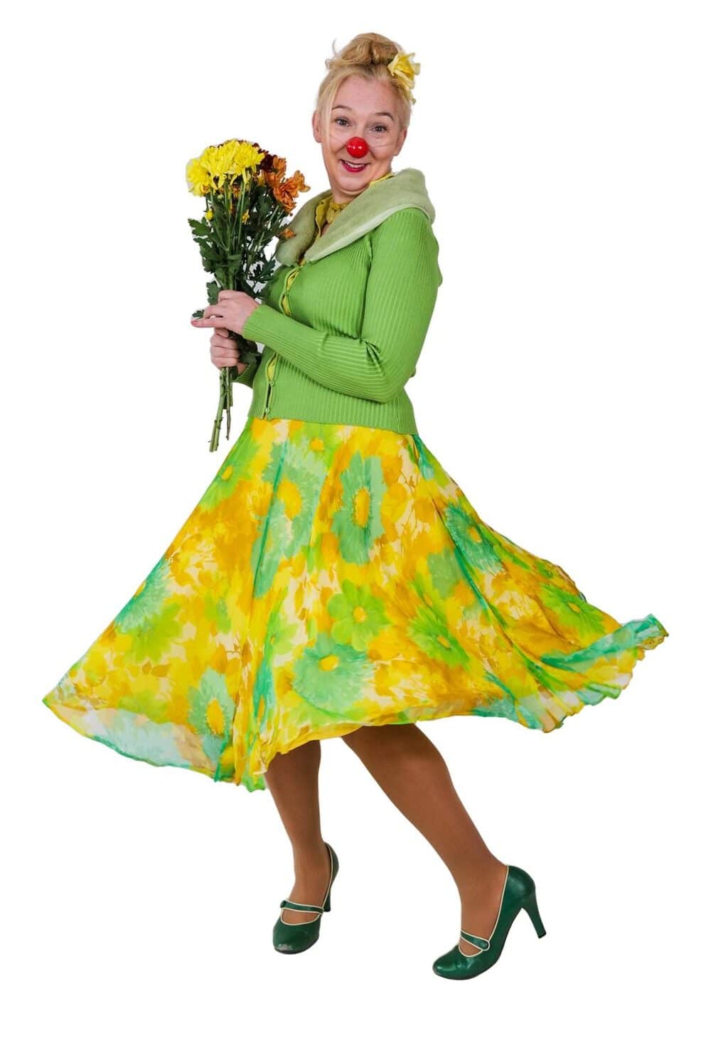 Portrait of a female clown with blonde hair, wearing a red nose, a green cardigan, a green and yellow floral skirt, green heels, and a yellow flower in her hair, holding a bouquet of yellow and brown flowers.