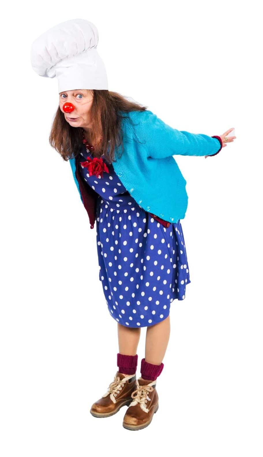 Portrait of a female clown wearing a white chef's hat, a red nose, a blue polka dot dress, and a blue cardigan, leaning forward with a curious expression.