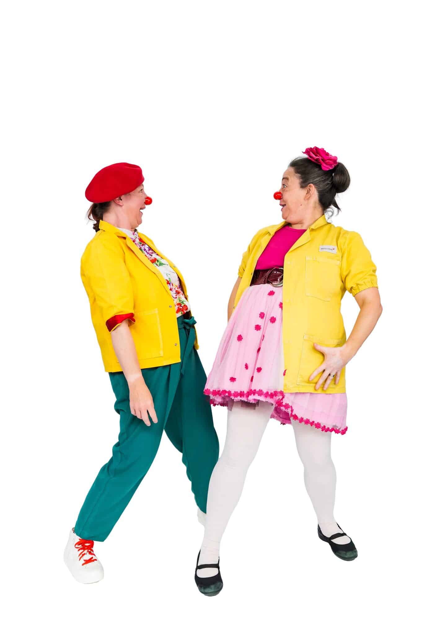 Two female clowns standing side by side laughing together. Both wear yellow jackets and red noses; the one on the left wears a red beret and teal pants, while the one on the right wears a pink tutu skirt and has a pink flower in her hair.
