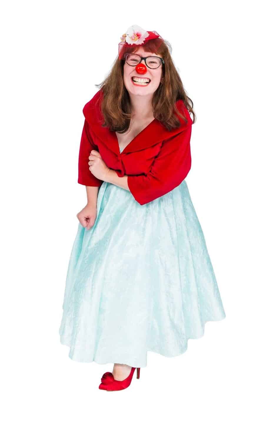 Portrait of a smiling female clown wearing glasses, a red nose, a red velvet bolero jacket, a light blue skirt, red heels, and a flower in her hair.